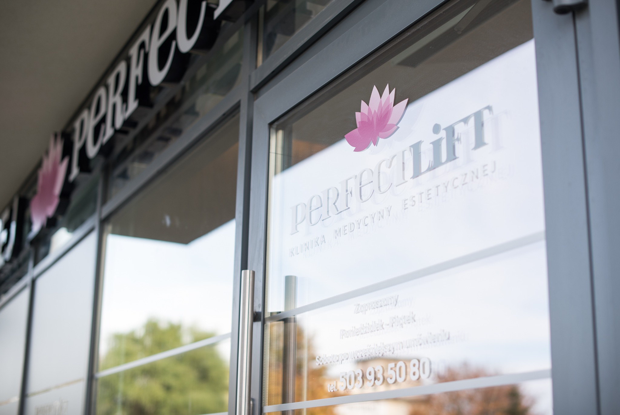 Wejście do kliniki medycyny estetycznej Perfect Lift z logo kwiatu lotosu na szybie i numerem telefonu.