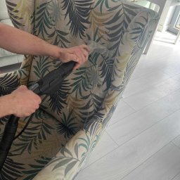 Clean Secure Duo - Czyszczenie ekstrakcyjne fotela z wzorem liści. Widoczne ręce osoby czyszczącej i dysza odkurzacza piorącego. Jasna podłoga i fragment pokoju w tle.