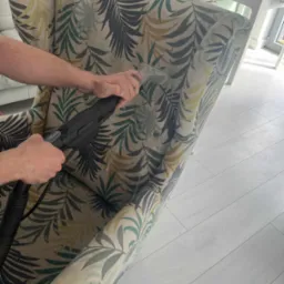 Czyszczenie ekstrakcyjne fotela z wzorem liści. Widoczne ręce osoby czyszczącej i dysza odkurzacza piorącego. Jasna podłoga i fragment pokoju w tle.