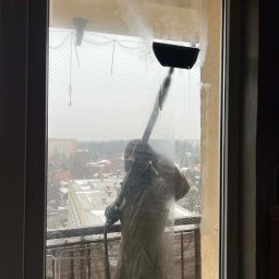 Clean Secure Duo - Mycie okien na balkonie wysokociśnieniową myjką. Widok na miasto przez siatkę zabezpieczającą. Częściowo umyte okno z zaciekami. Osoba w kombinezonie ochronnym.