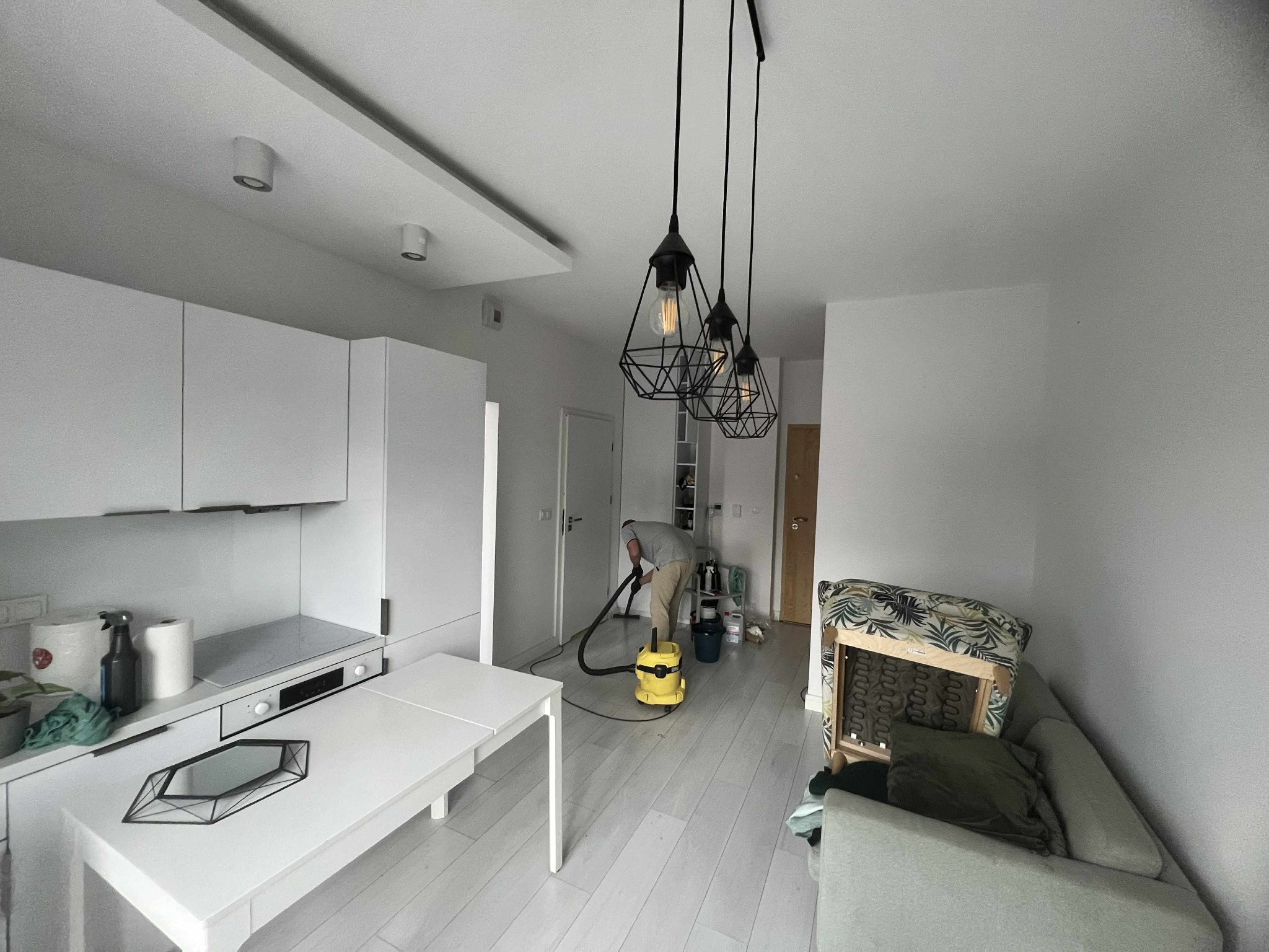 Mężczyzna odkurza podłogę w jasnym wnętrzu z aneksem kuchennym i designerskimi lampami. Widoczny sprzęt czyszczący i częściowo rozebrana sofa.