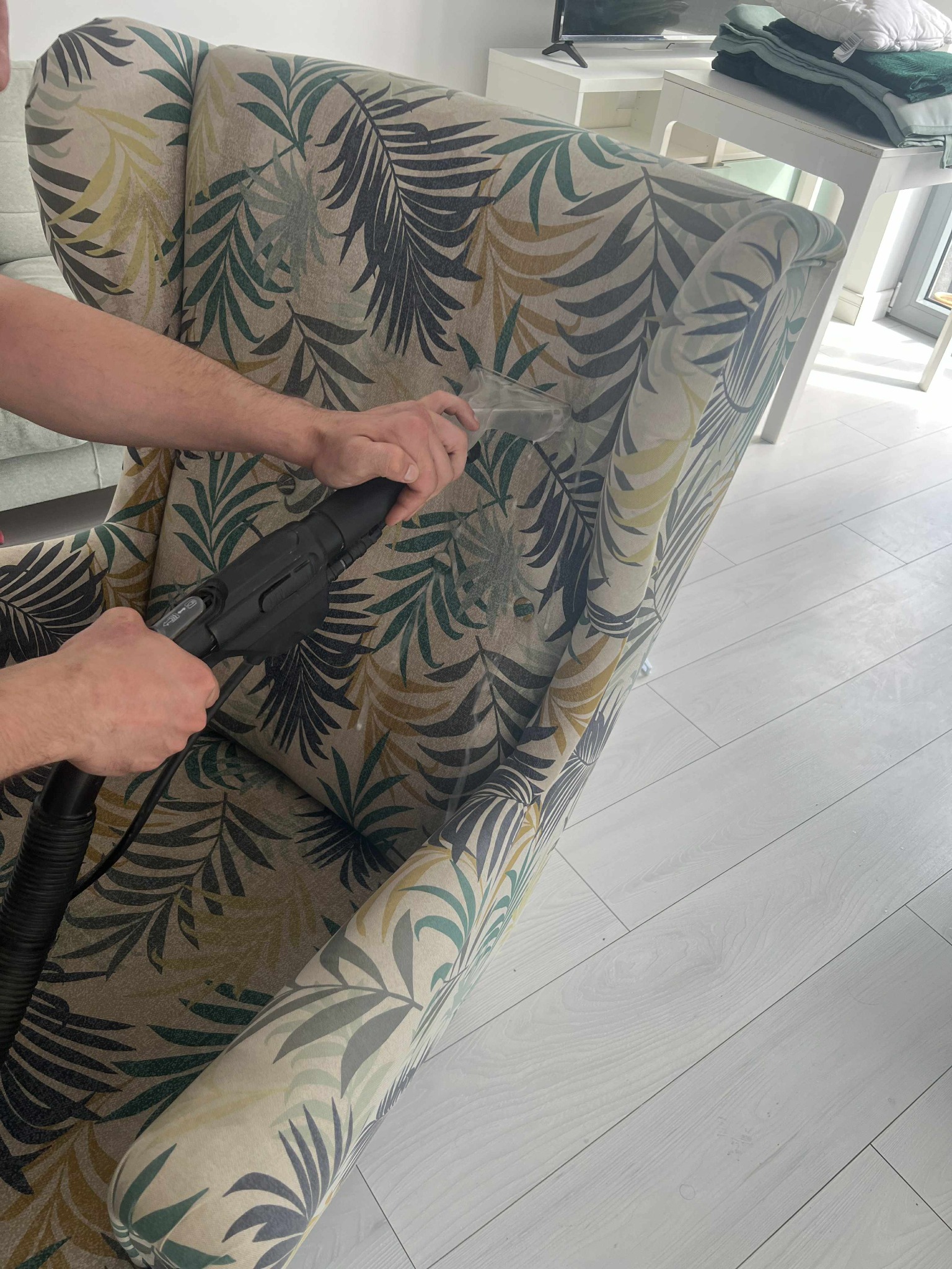 Czyszczenie ekstrakcyjne fotela z wzorem liści. Widoczne ręce osoby czyszczącej i dysza odkurzacza piorącego. Jasna podłoga i fragment pokoju w tle.
