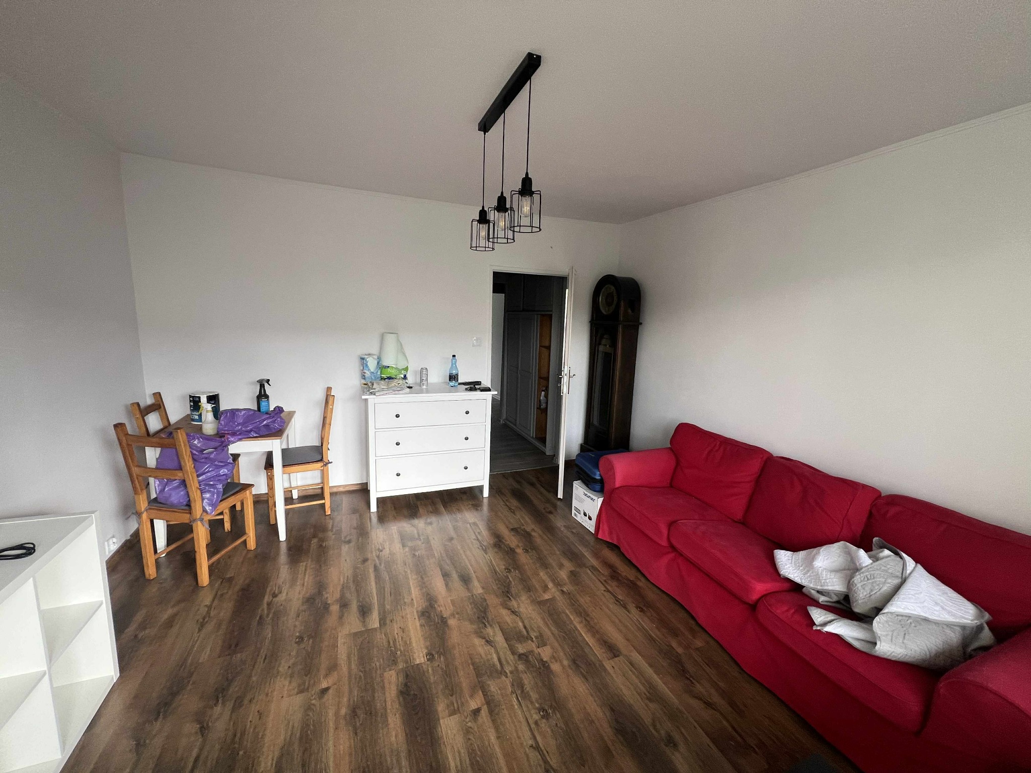 Jasne wnętrze pokoju z drewnianą podłogą, czerwona sofa i białe meble. Stolik przy ścianie z widocznymi środkami czystości. Widok na odnowione mieszkanie.