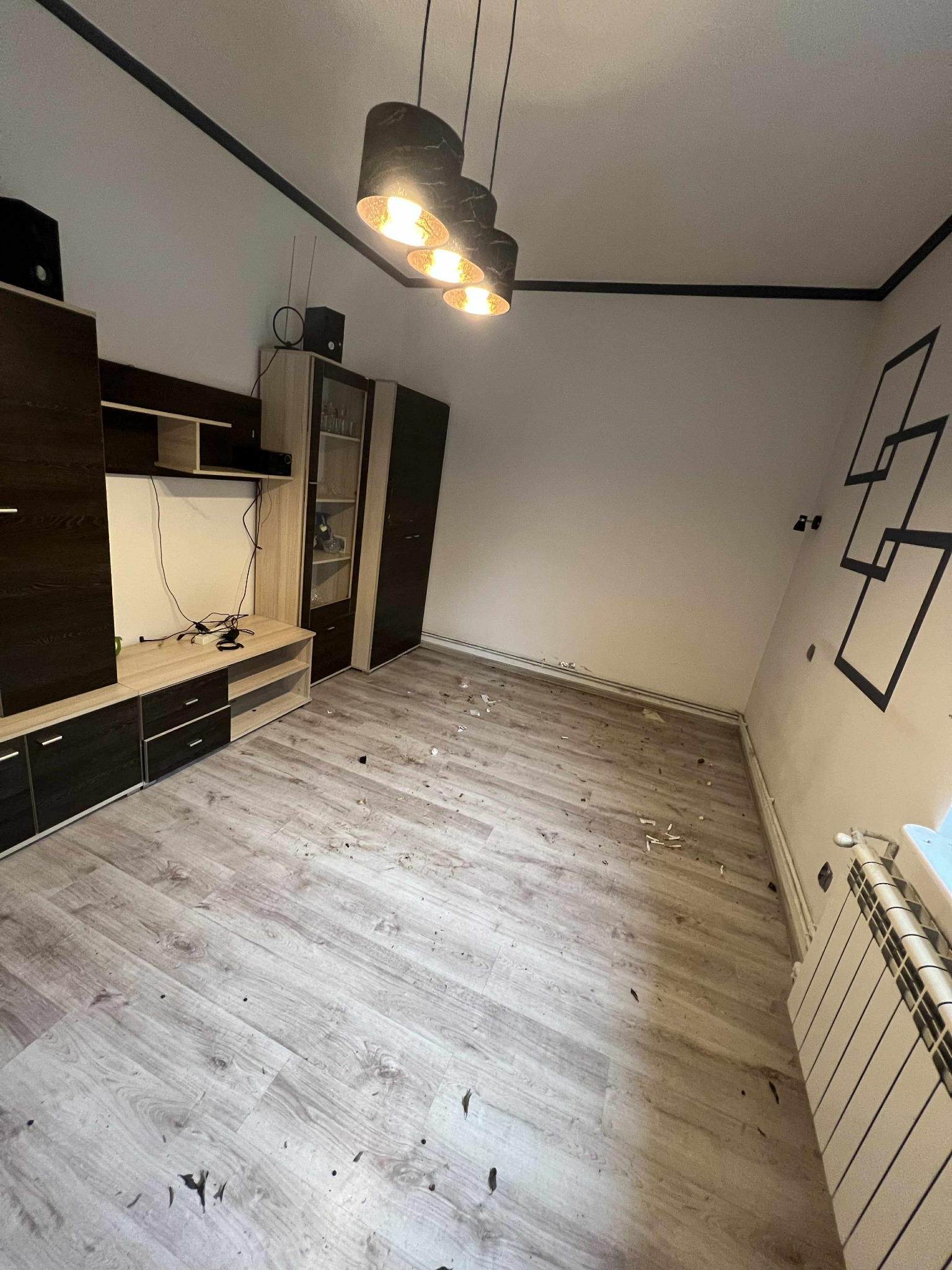 Pokój po remoncie z rozsypanymi resztkami gruzu na podłodze. Widoczny regał, lampa i grzejnik. Na ścianie dekoracyjne, czarne kwadraty.