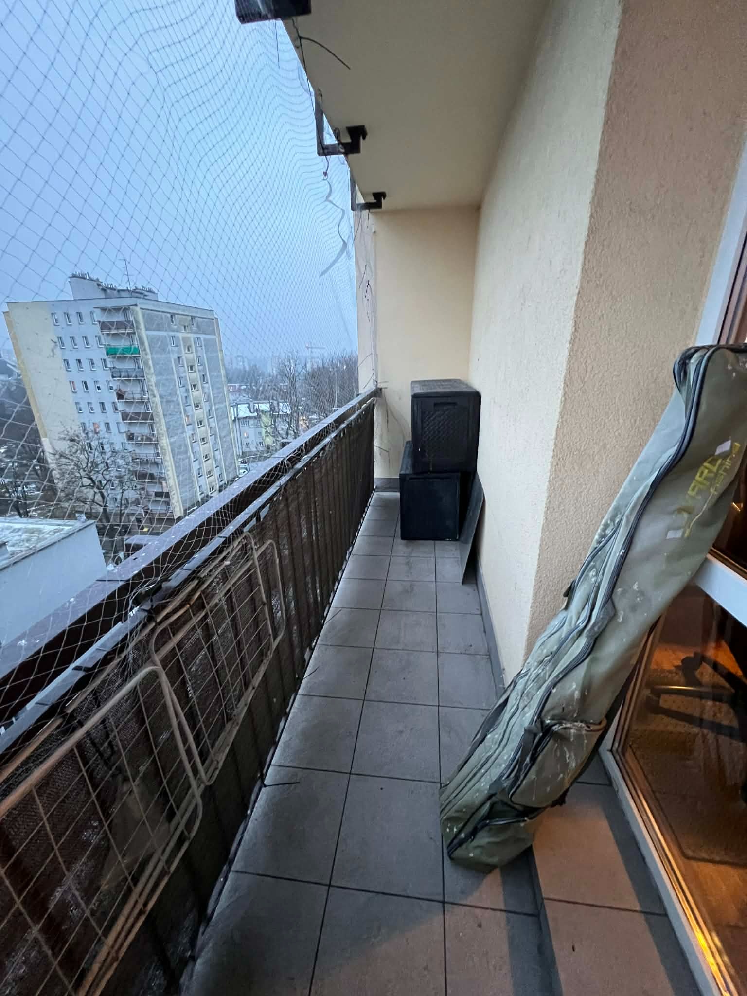 Zabezpieczony siatką balkon z widokiem na blok. Na podłodze skrzynki i pokrowiec na wędki. Szara kolorystyka, pochmurny dzień. Widok z góry.