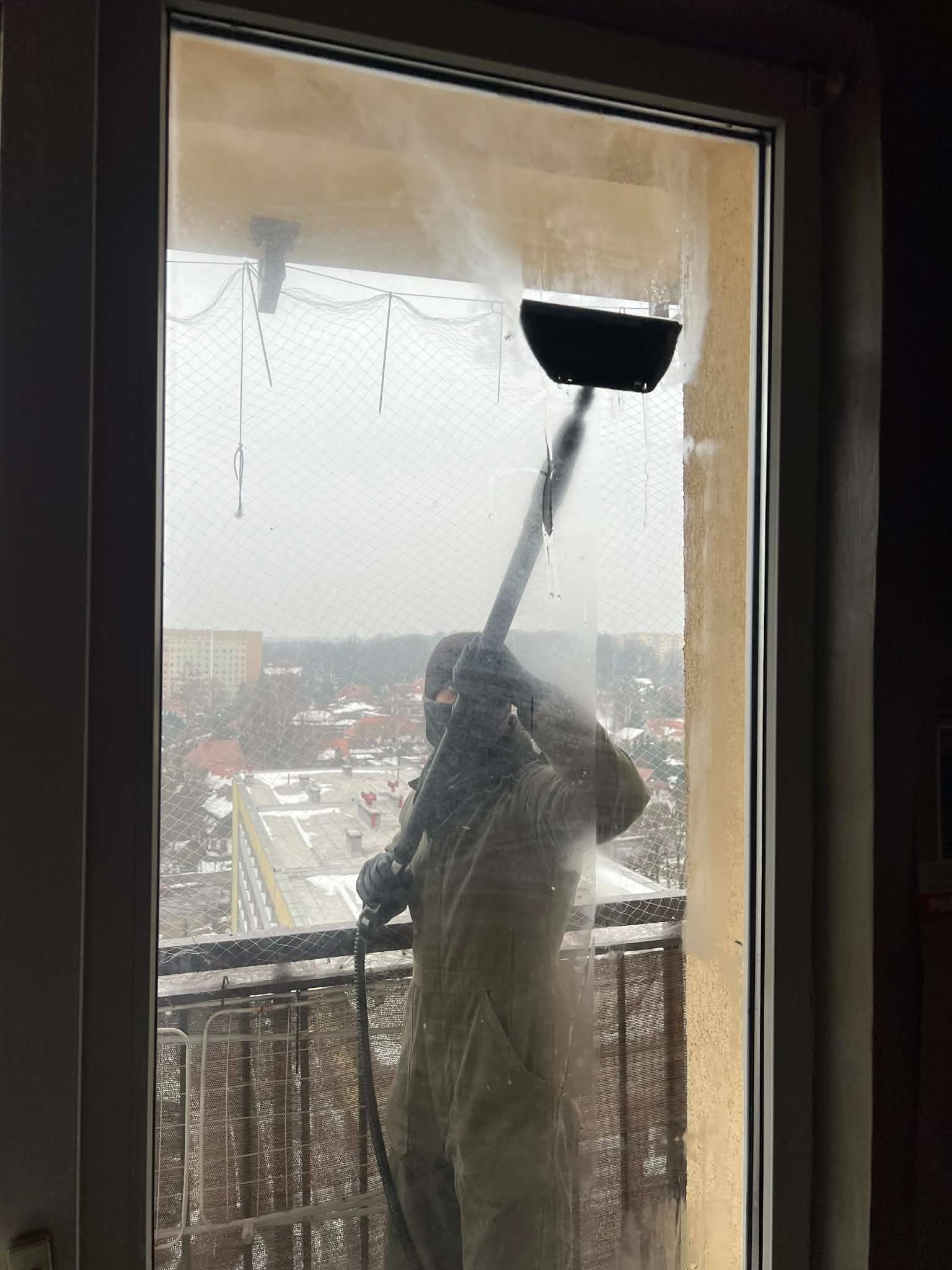 Mycie okien na balkonie wysokociśnieniową myjką. Widok na miasto przez siatkę zabezpieczającą. Częściowo umyte okno z zaciekami. Osoba w kombinezonie ochronnym.