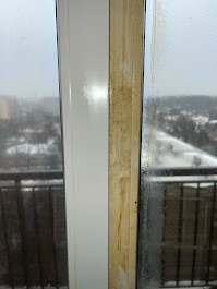 Okno balkonowe: połowa ramy i szyby wyczyszczona, druga brudna. Widok na miasto w tle, pochmurny dzień. Kontrast czystości i zabrudzeń.