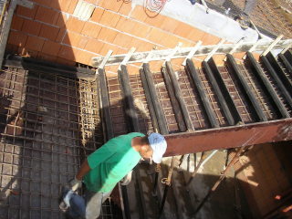 Pracownik w zielonej koszulce i czapce montuje zbrojenie pod posadzkę jastrychową wewnątrz budynku w trakcie budowy. Widoczne szalunki i ceglany mur.