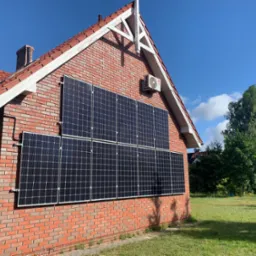 Czerwony ceglany dom z panelami słonecznymi na ścianie i klimatyzatorem LG pod okapem dachu, widok z zewnątrz, błękitne niebo.
