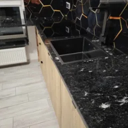 Czarny blat kuchenny z granitu z widocznymi białymi żyłkami, zintegrowany z ciemnym zlewem i drewnianymi szafkami; w tle ściana z heksagonalnymi płytkami z podświetleniem.