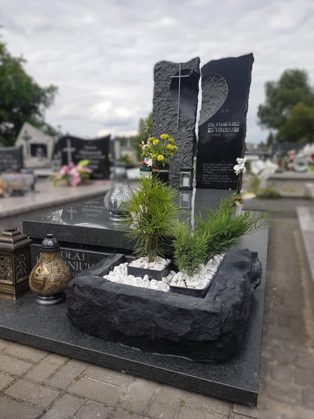 Czarny, granitowy pomnik z dwoma pionowymi płytami, jedna z wyciętym krzyżem, druga z inskrypcją, ozdobiony kwiatami i roślinami w kamiennej donicy z białymi kamykami, na tle cmentarza.