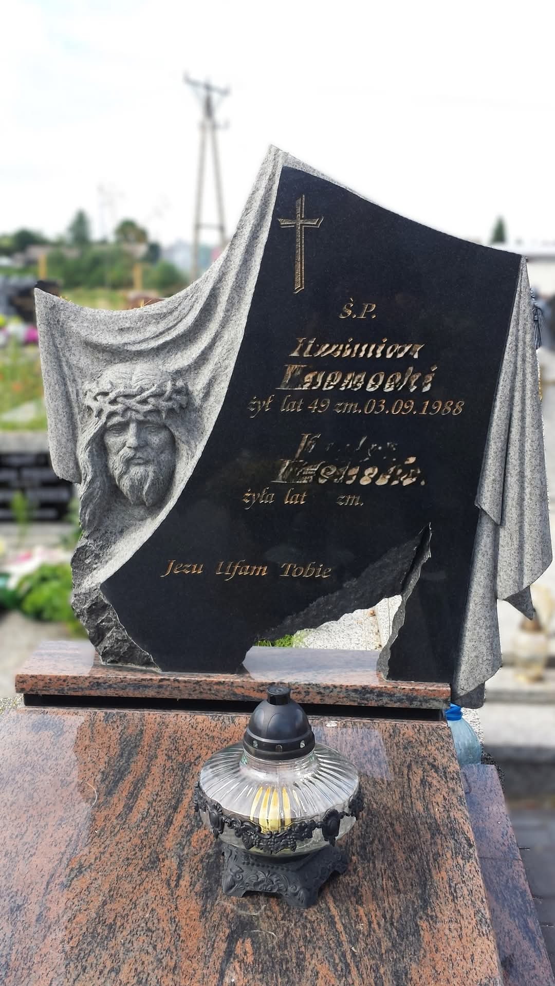 Czarny nagrobek z rzeźbioną postacią Jezusa w koronie cierniowej i napisem 'Jezu Ufam Tobie', ustawiony na podstawie z czerwonego granitu z zapalonym zniczem.