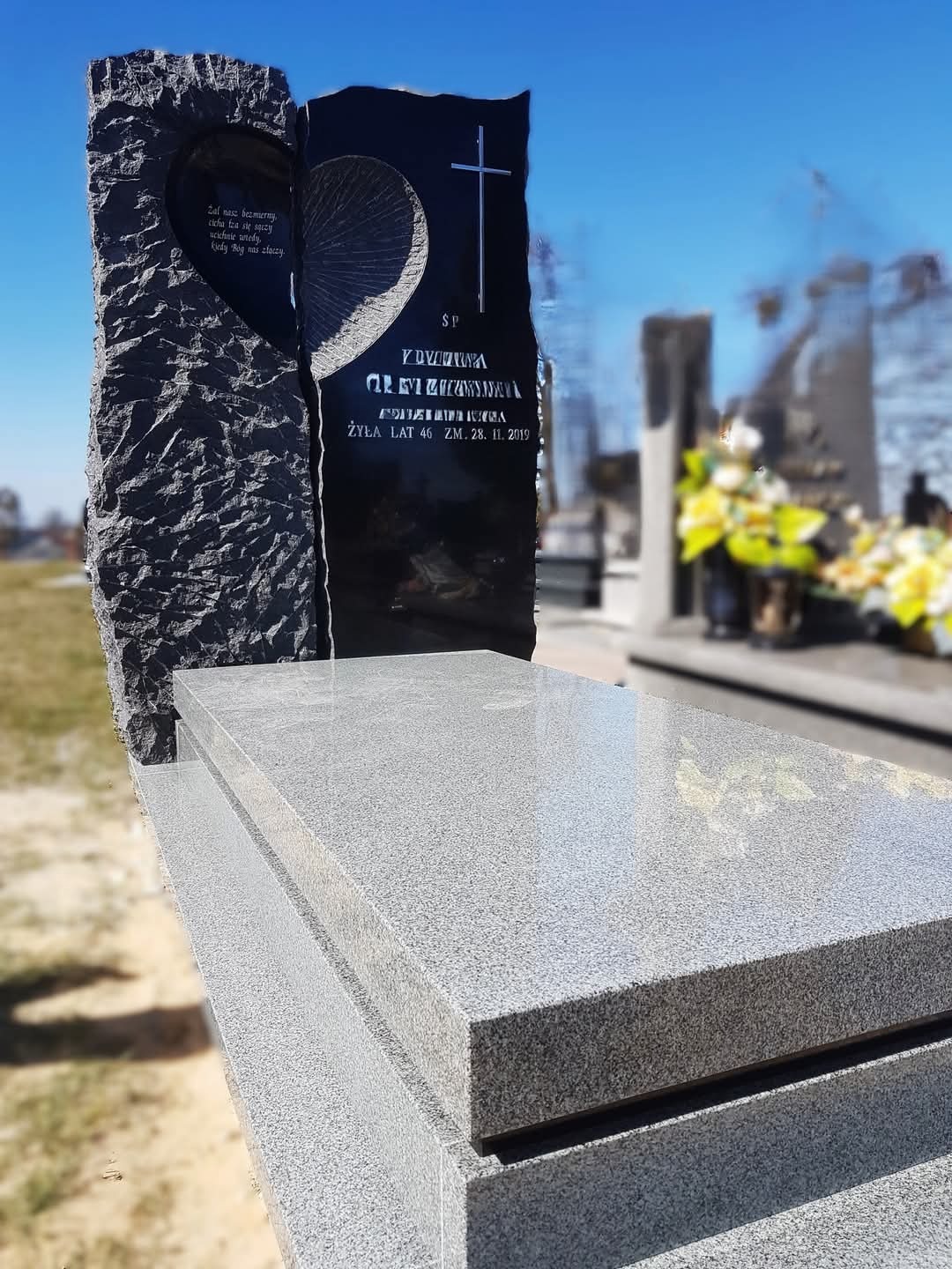 Granitowy pomnik z dwoma pionowymi płytami: jedna z surową fakturą i wyciętym sercem, druga gładka z krzyżem i inskrypcją, na jasnoszarym grobie, w tle inne nagrobki i kwiaty.