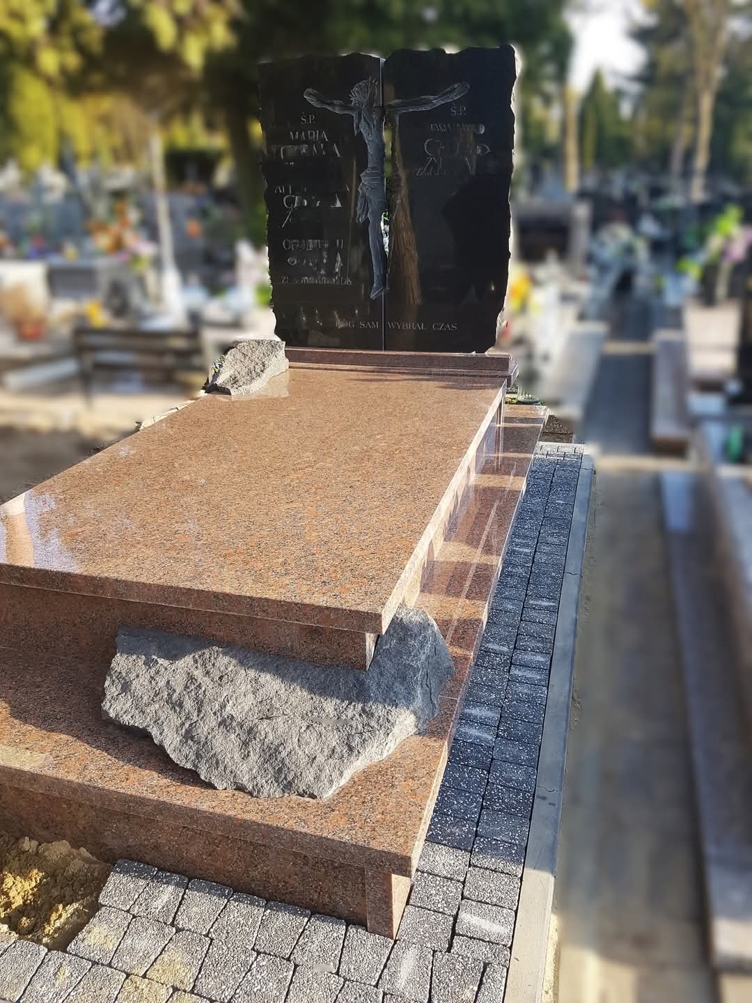 Granitowy pomnik nagrobny z ciemną, rzeźbioną płytą przedstawiającą ukrzyżowanie, osadzony na stopniowanej podstawie z jasnego granitu, otoczony kostką brukową.