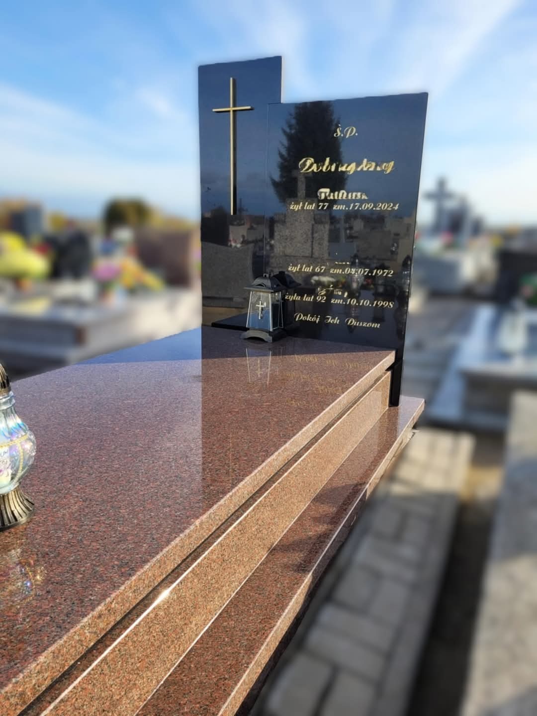 Granitowy pomnik z ciemną tablicą z wygrawerowanym złotym krzyżem i inskrypcjami, z lampionem i kwiatami w tle cmentarza.