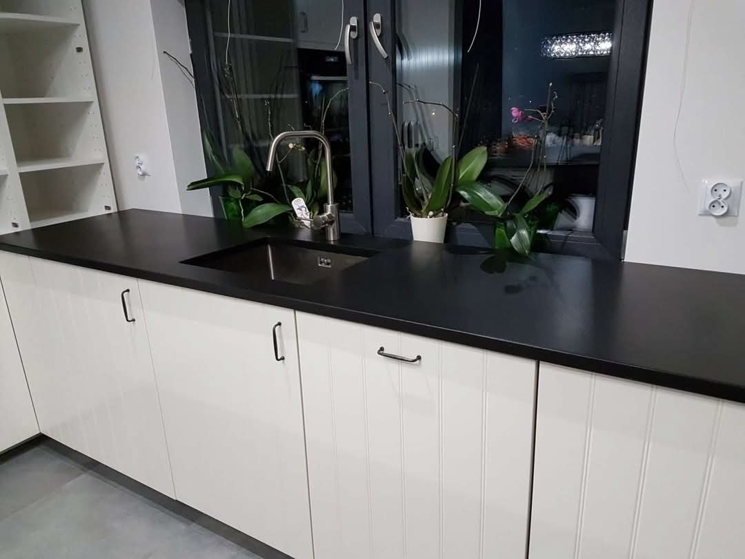 Czarny blat kamienny z wpuszczonym zlewem i baterią, białe szafki kuchenne z podłużnymi frezowaniami, roślina w doniczce na tle okna.