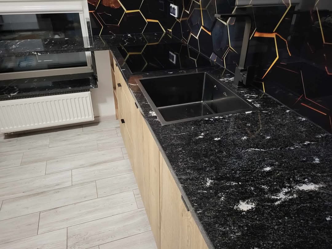 Czarny blat kuchenny z granitu z widocznymi białymi żyłkami, zintegrowany z ciemnym zlewem i drewnianymi szafkami; w tle ściana z heksagonalnymi płytkami z podświetleniem.