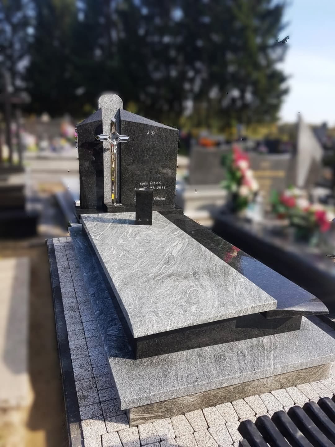 Czarny granitowy pomnik z srebrnym krzyżem i szarą płytą nagrobkową o marmurkowej fakturze, na cmentarzu.