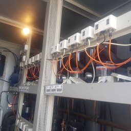 FRIGOPARTNER Bogdan Kobyliński - Szereg białych modułów Danfoss REG SPR.5 i ZABEZP SPR.x z pomarańczowymi kablami, zamontowanych na metalowej konstrukcji w pomieszczeniu technicznym, oświetlonych górną lampą.