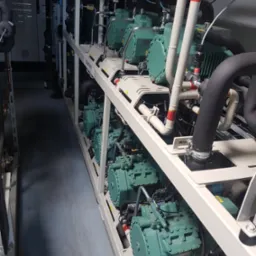 Rząd sprężarek chłodniczych w przemysłowej instalacji, z widocznymi rurami, zaworami i elementami sterującymi, umieszczonych na metalowej ramie w pomieszczeniu technicznym.