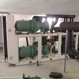 FRIGOPARTNER Bogdan Kobyliński - Agregat chłodniczy przemysłowy z dwoma sprężarkami w pomieszczeniu technicznym, widoczne rury izolowane termicznie i skrzynki narzędziowe na posadzce.