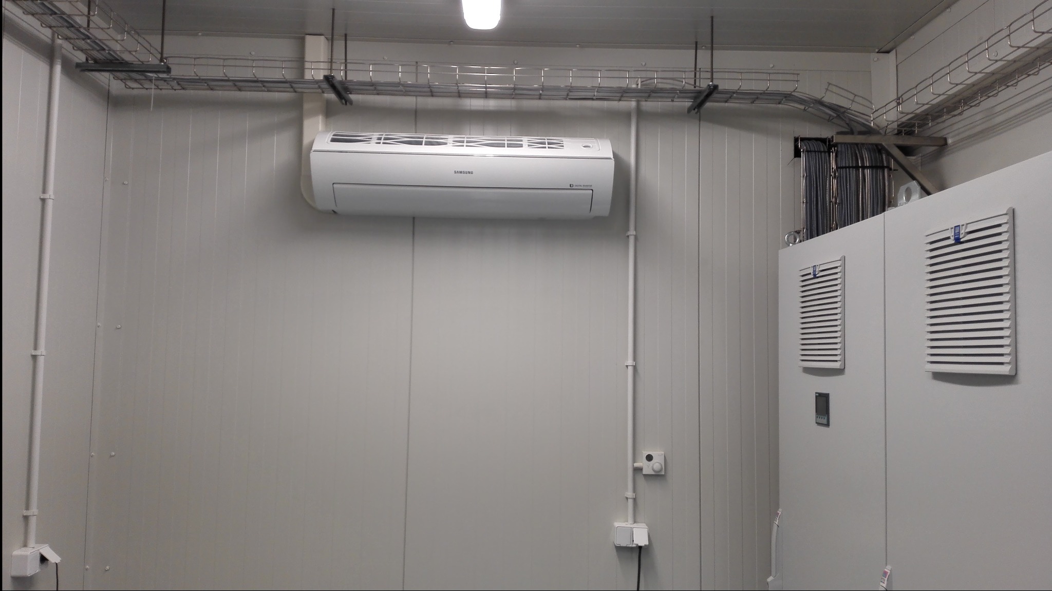Wewnętrzna jednostka klimatyzacji Samsung zamontowana na panelowej ścianie w pomieszczeniu technicznym, widoczne koryta kablowe pod sufitem i szafa sterownicza z kratkami wentylacyjnymi.