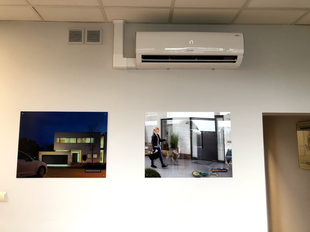 Biała klimatyzacja ścienna z wyświetlaczem temperatury 17 stopni, zamontowana na ścianie nad dwoma obrazami reklamowymi firmy Hormann.