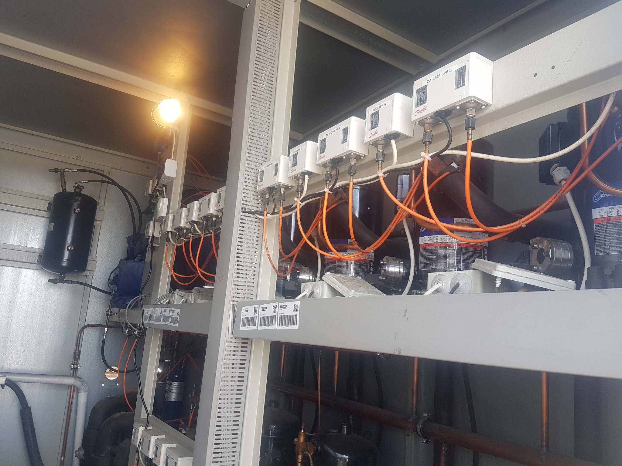 Szereg białych modułów Danfoss REG SPR.5 i ZABEZP SPR.x z pomarańczowymi kablami, zamontowanych na metalowej konstrukcji w pomieszczeniu technicznym, oświetlonych górną lampą.