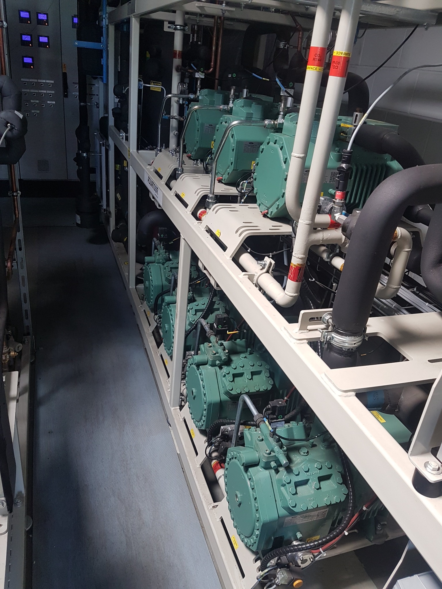 Rząd sprężarek chłodniczych w przemysłowej instalacji, z widocznymi rurami, zaworami i elementami sterującymi, umieszczonych na metalowej ramie w pomieszczeniu technicznym.