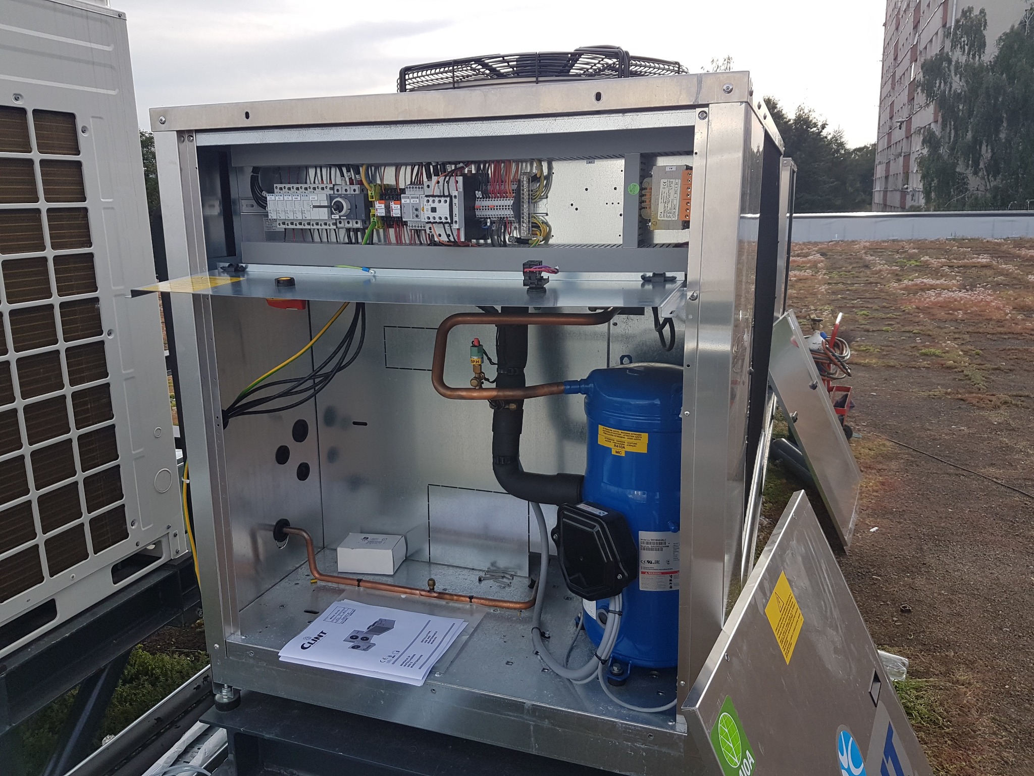 Otwarta obudowa agregatu chłodniczego na dachu budynku, widoczne elementy instalacji elektrycznej, rury miedziane, kompresor w kolorze niebieskim oraz dokumentacja techniczna Clint.