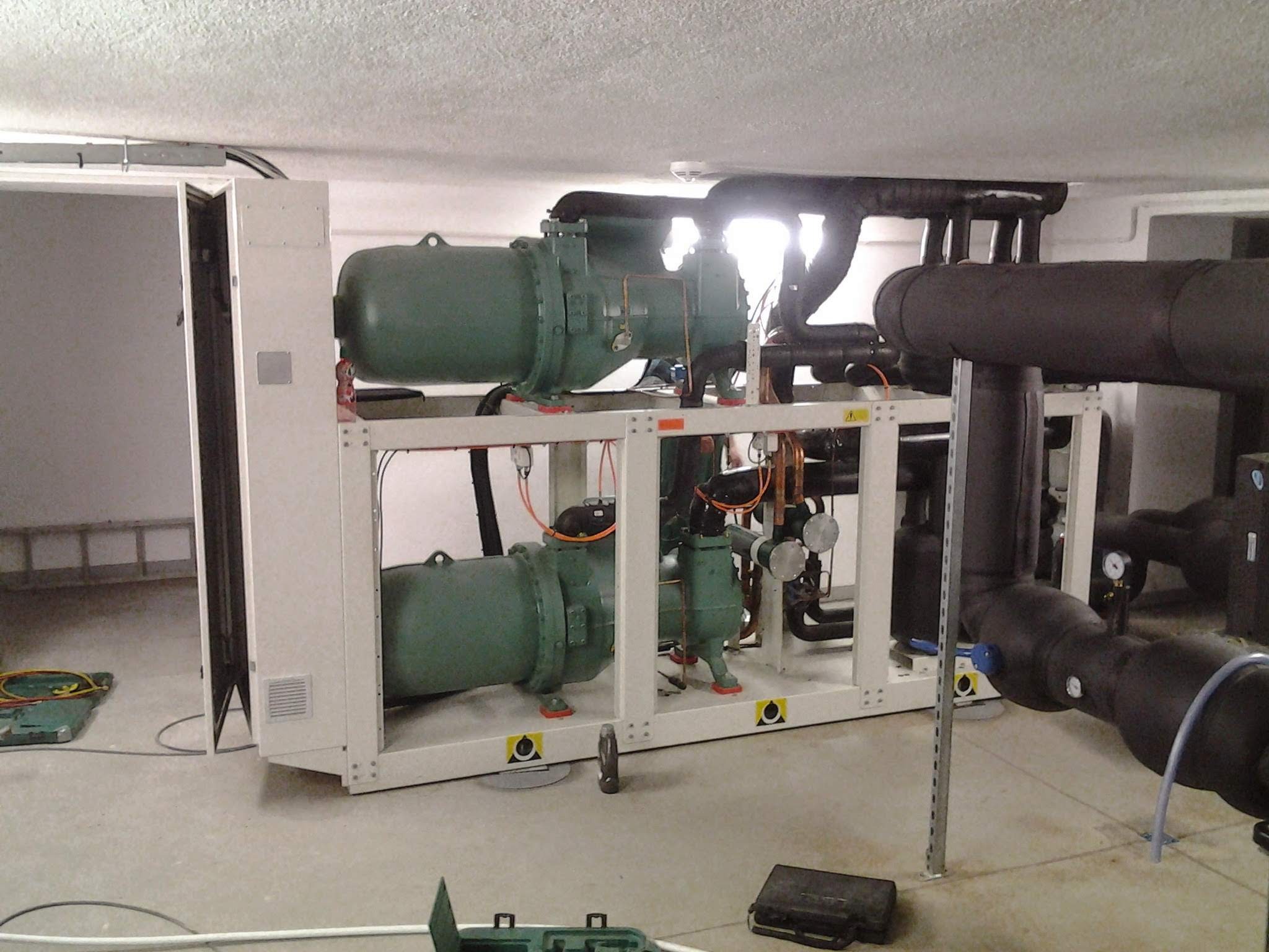 Agregat chłodniczy przemysłowy z dwoma sprężarkami w pomieszczeniu technicznym, widoczne rury izolowane termicznie i skrzynki narzędziowe na posadzce.