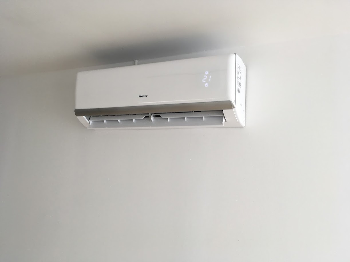 Biała klimatyzacja marki Gree zamontowana na jasnej ścianie, wyświetlająca temperaturę 21 stopni Celsjusza.
