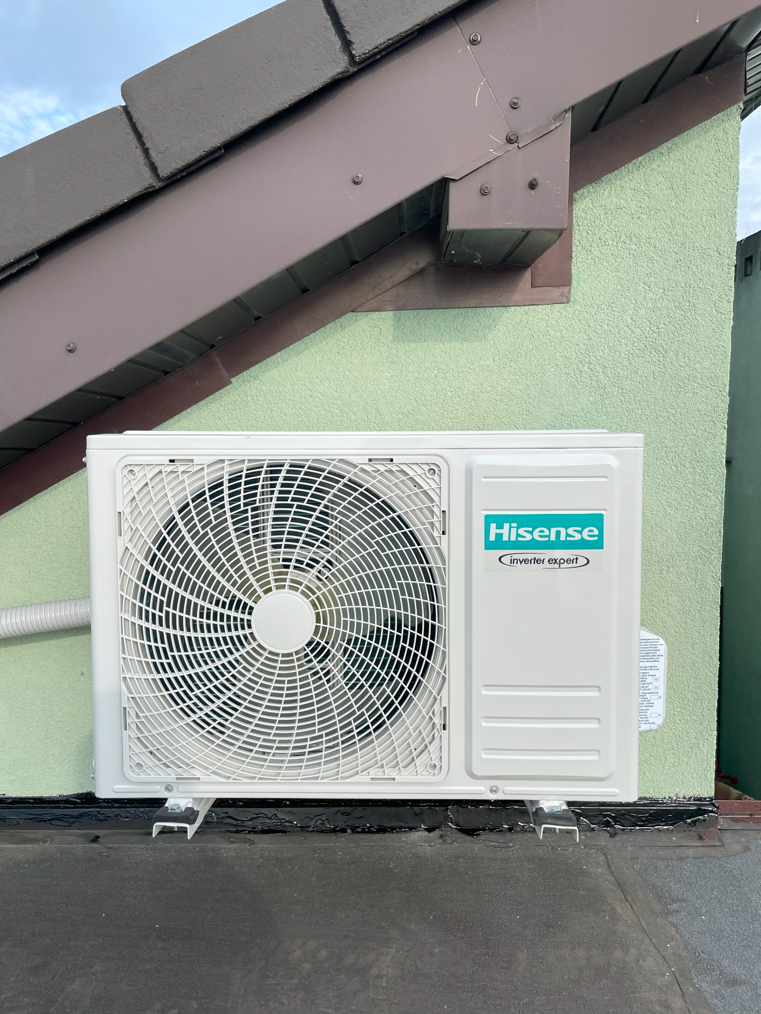 Biała jednostka zewnętrzna klimatyzacji Hisense zamontowana na dachu budynku z zieloną elewacją i brązowym okapem, widoczny przewód.