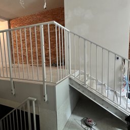 Artur Prokofiev - Białe, metalowe balustrady schodowe w surowym wnętrzu z ceglaną ścianą w tle. Widoczne narzędzia i kable, sugerujące trwający montaż.