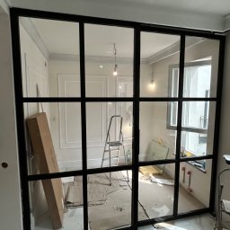 Artur Prokofiev - Przeszklona ścianka działowa z czarnymi szprosami w Szczecinie. Widoczne kartony i drabina, sugerujące montaż w trakcie wykończenia wnętrza.