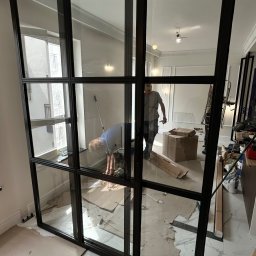 Artur Prokofiev - Montaż nowoczesnej, przesuwnej ścianki działowej z czarnymi profilami i szklanymi panelami. Widoczni montażyści podczas pracy, zabezpieczone podłoże.