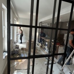 Artur Prokofiev - Loftowe przesuwne drzwi z czarną metalową ramą i szklanymi panelami oddzielają pomieszczenia. W tle widoczny malarz przy pracy.