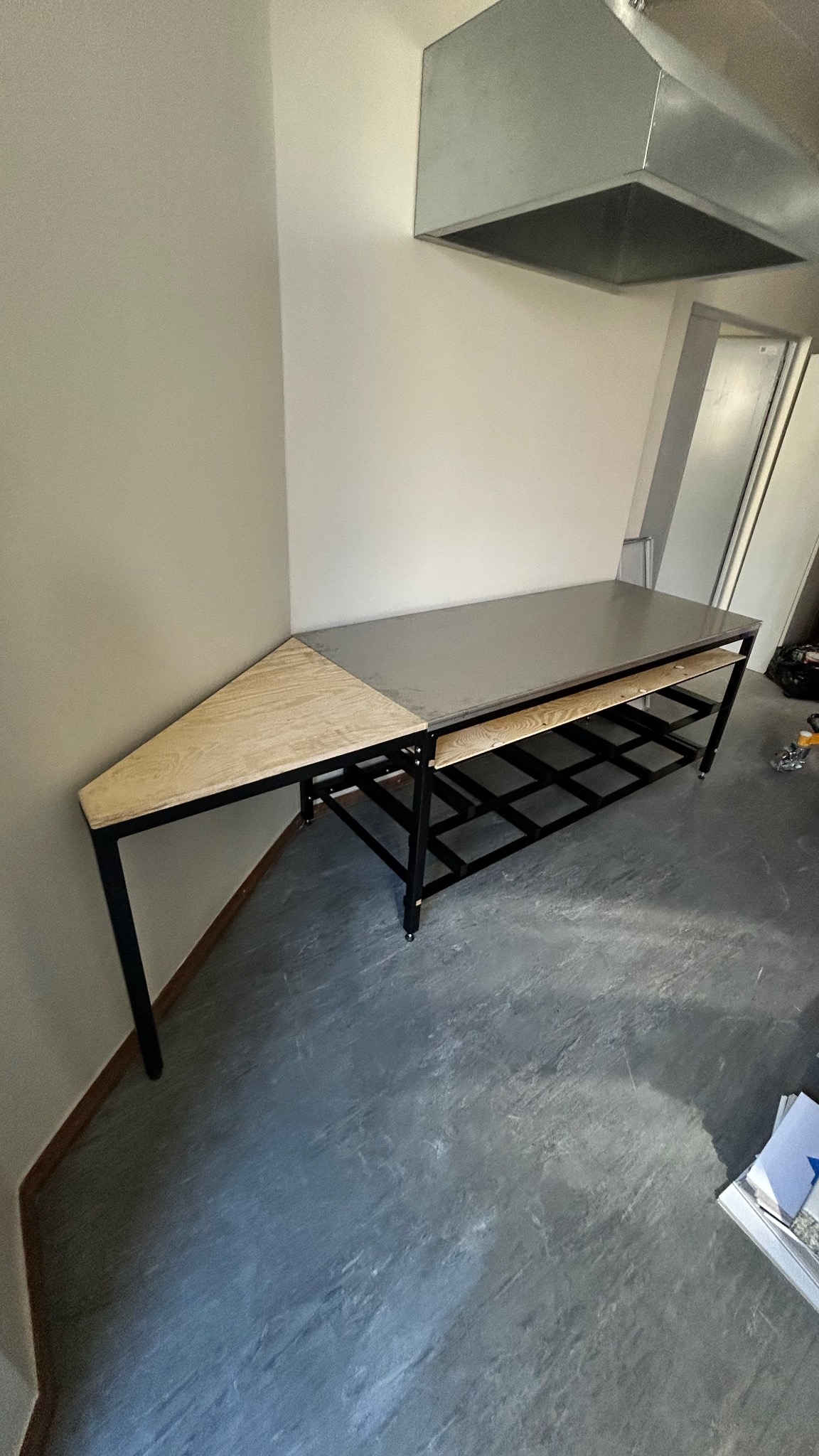 Stół roboczy z metalowym blatem i drewnianym narożnikiem, czarna metalowa konstrukcja, na kółkach, pod okapem. Surowe wykończenie, szara podłoga.