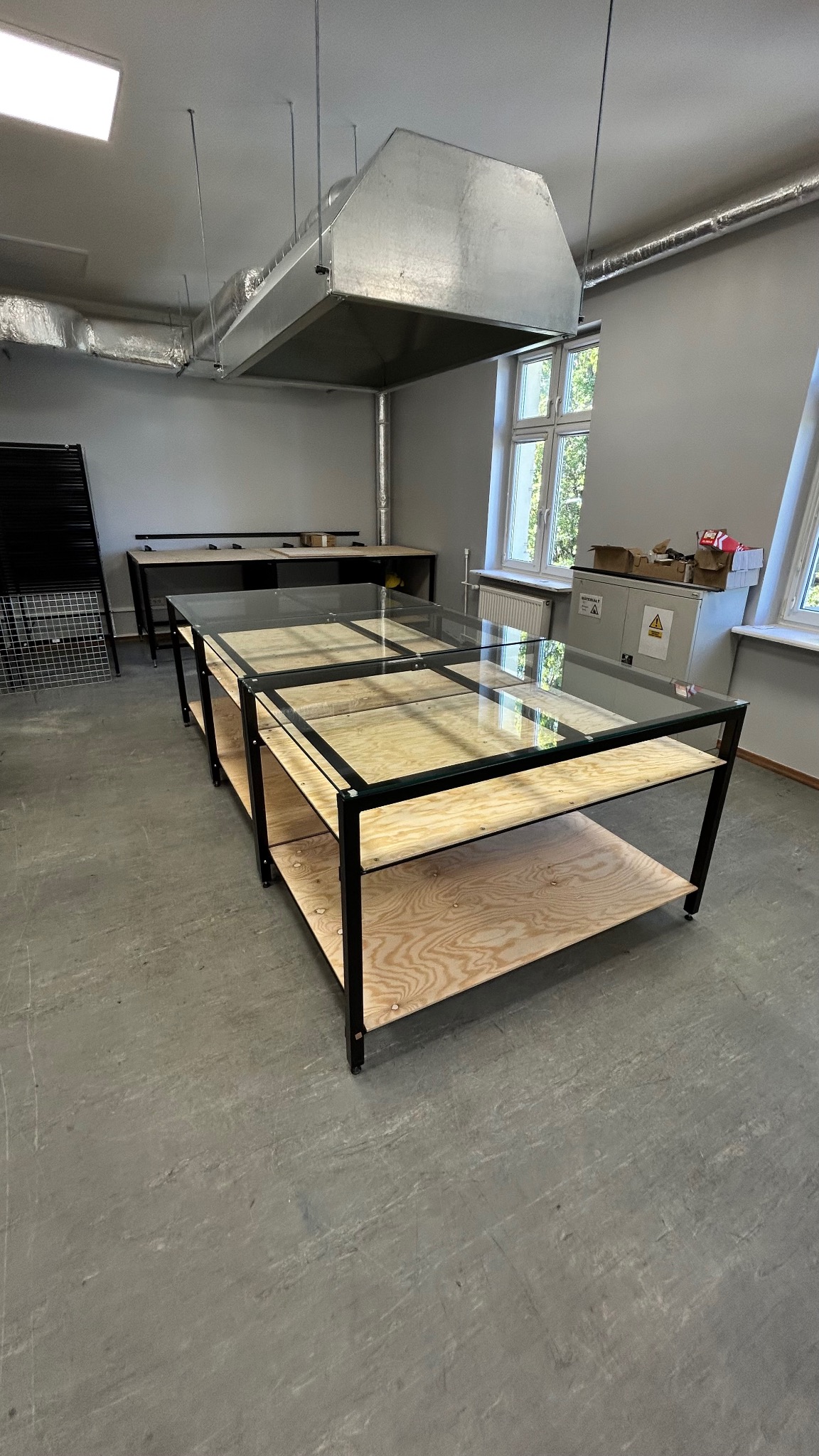 Metalowa konstrukcja stołów z szklanymi blatami i drewnianymi półkami w hali produkcyjnej. Widoczna wentylacja i okna.