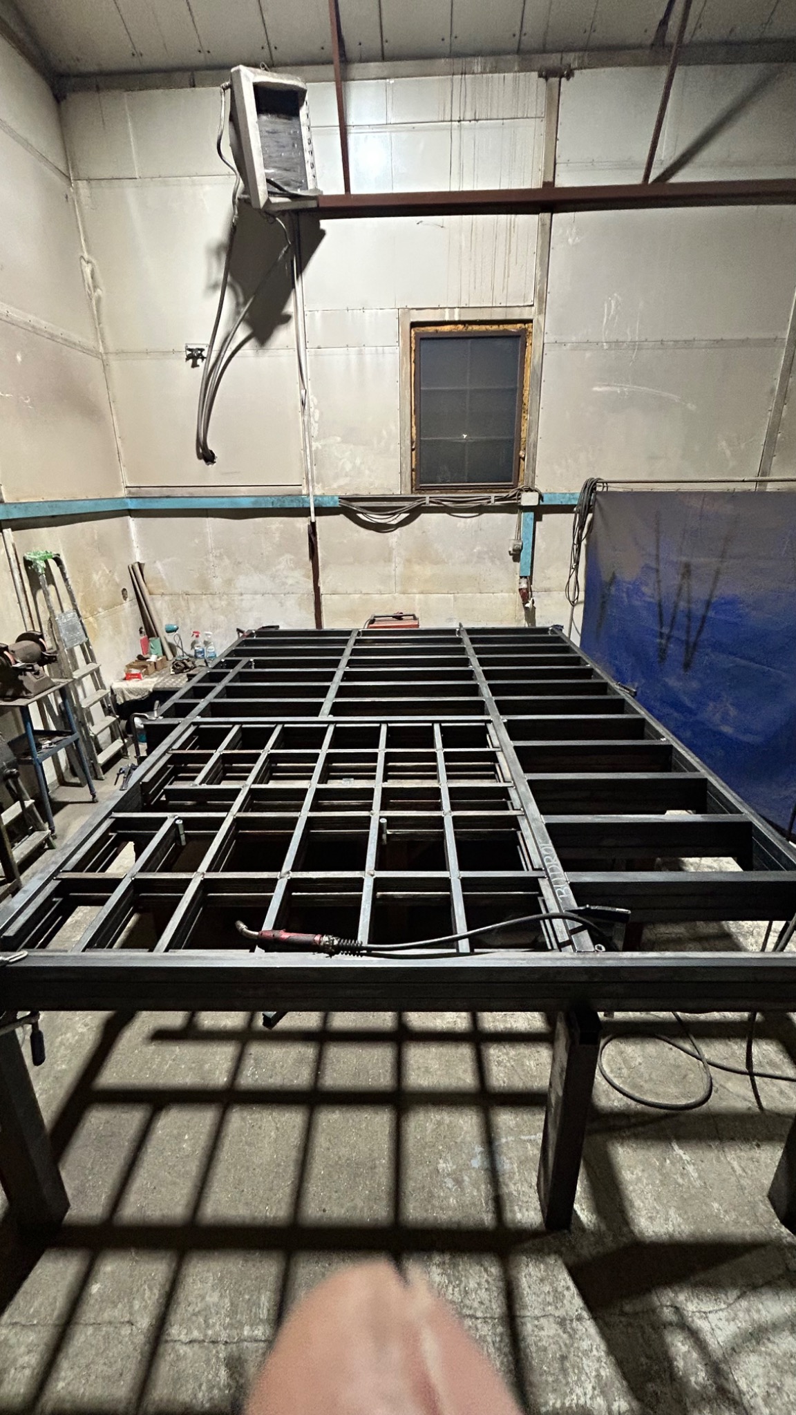 Metalowa konstrukcja kratowa w warsztacie, spawana z profili, widoczna spawarka, oświetlenie warsztatowe, surowe wnętrze hali.