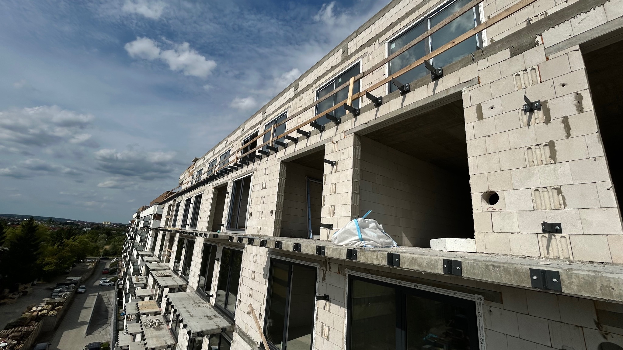 Wznoszenie wielopiętrowego budynku z białych bloków, widoczne okna i elementy konstrukcyjne balkonów, błękitne niebo z chmurami w tle. Surowy stan budynku.
