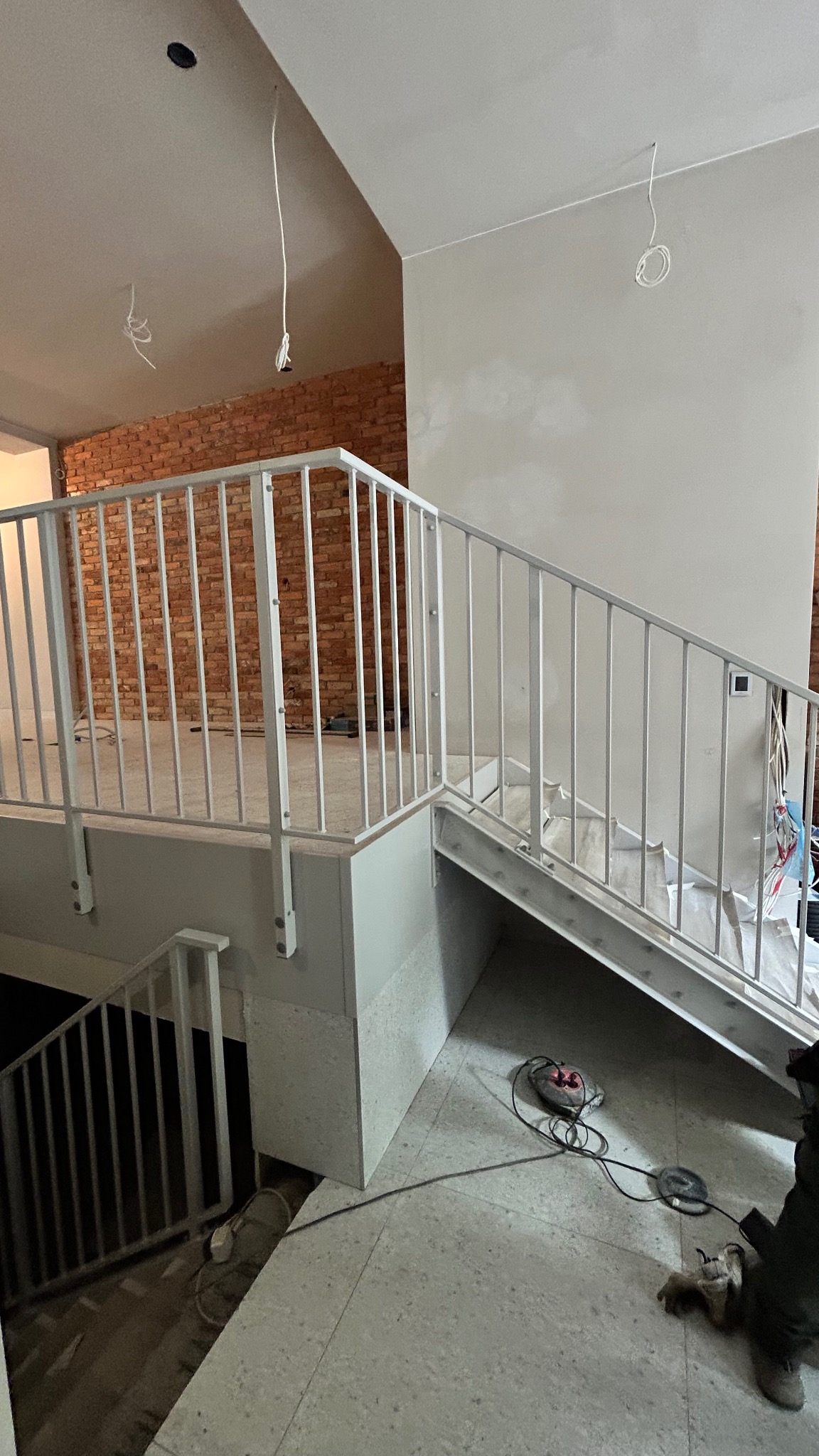 Białe, metalowe balustrady schodowe w surowym wnętrzu z ceglaną ścianą w tle. Widoczne narzędzia i kable, sugerujące trwający montaż.
