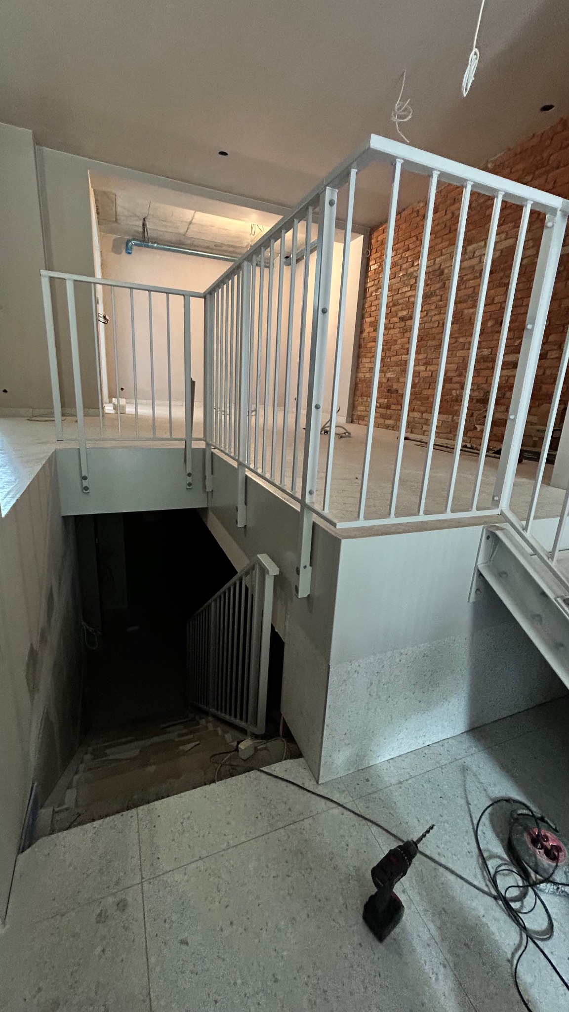Wnętrze w trakcie budowy z białą metalową balustradą schodową i poziomą. Widoczna ceglana ściana, w tle surowy beton. W pierwszym planie wiertarka.