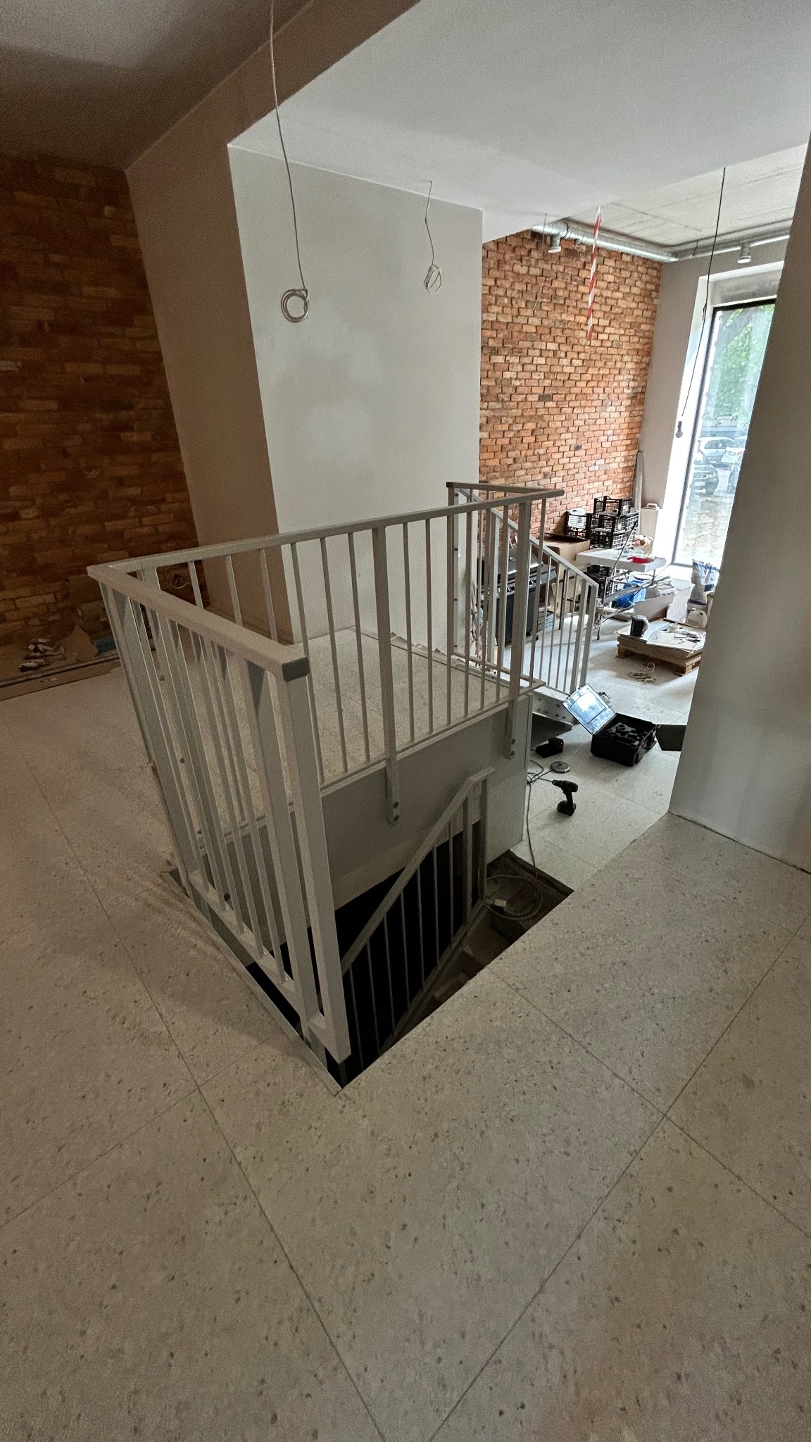 Biała metalowa balustrada schodowa z pionowymi prętami, widok z góry na schody w surowym wnętrzu z ceglaną ścianą i narzędziami.