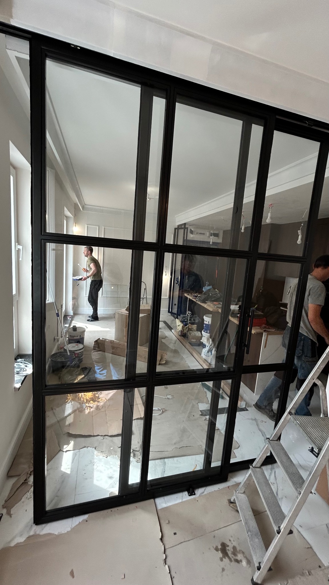 Loftowe przesuwne drzwi z czarną metalową ramą i szklanymi panelami oddzielają pomieszczenia. W tle widoczny malarz przy pracy.