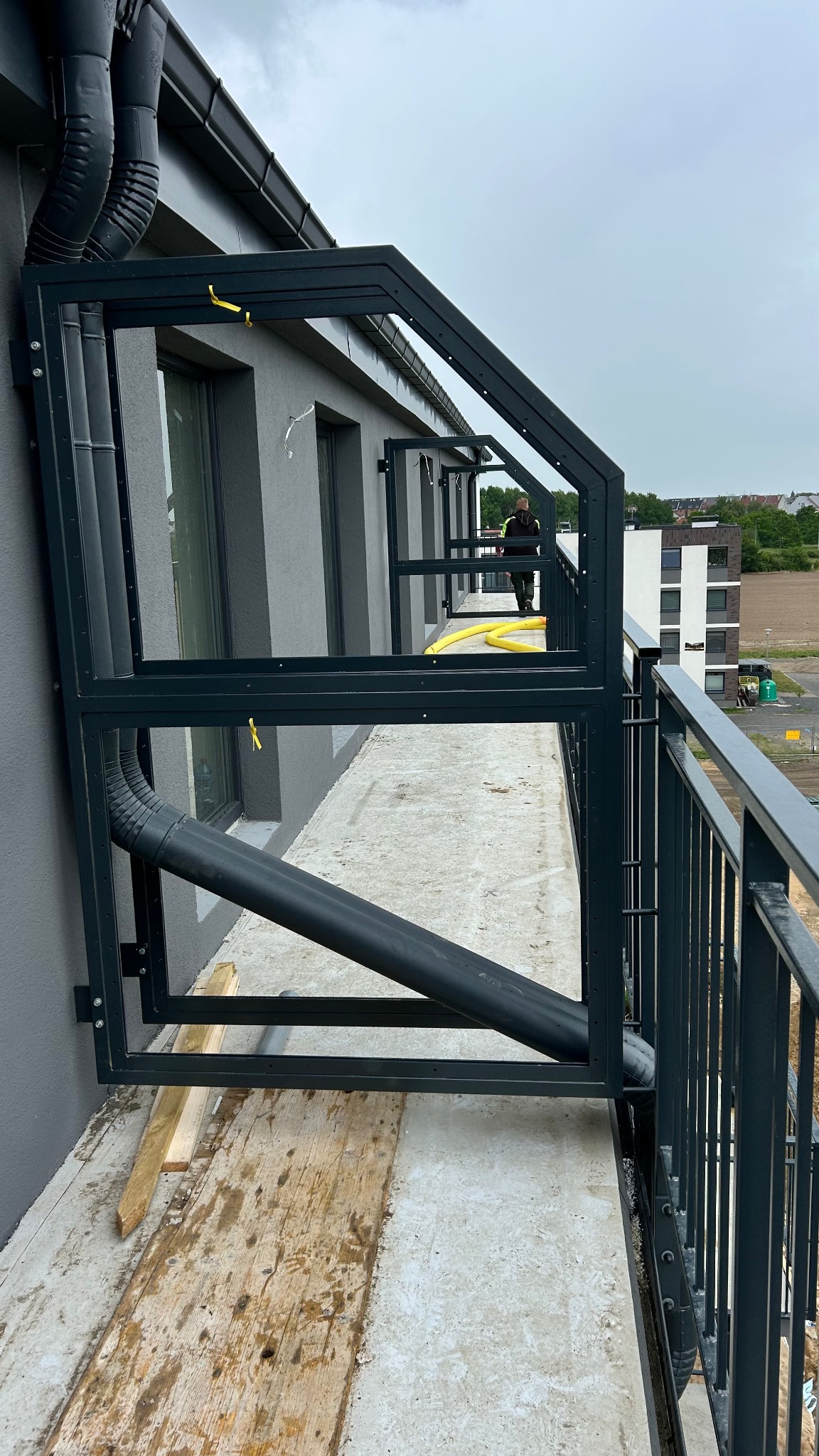 Montaż metalowej konstrukcji na balkonie budynku wielorodzinnego. Widoczne elementy mocujące, obróbka blacharska i fragment balustrady. Prace montażowe w toku.