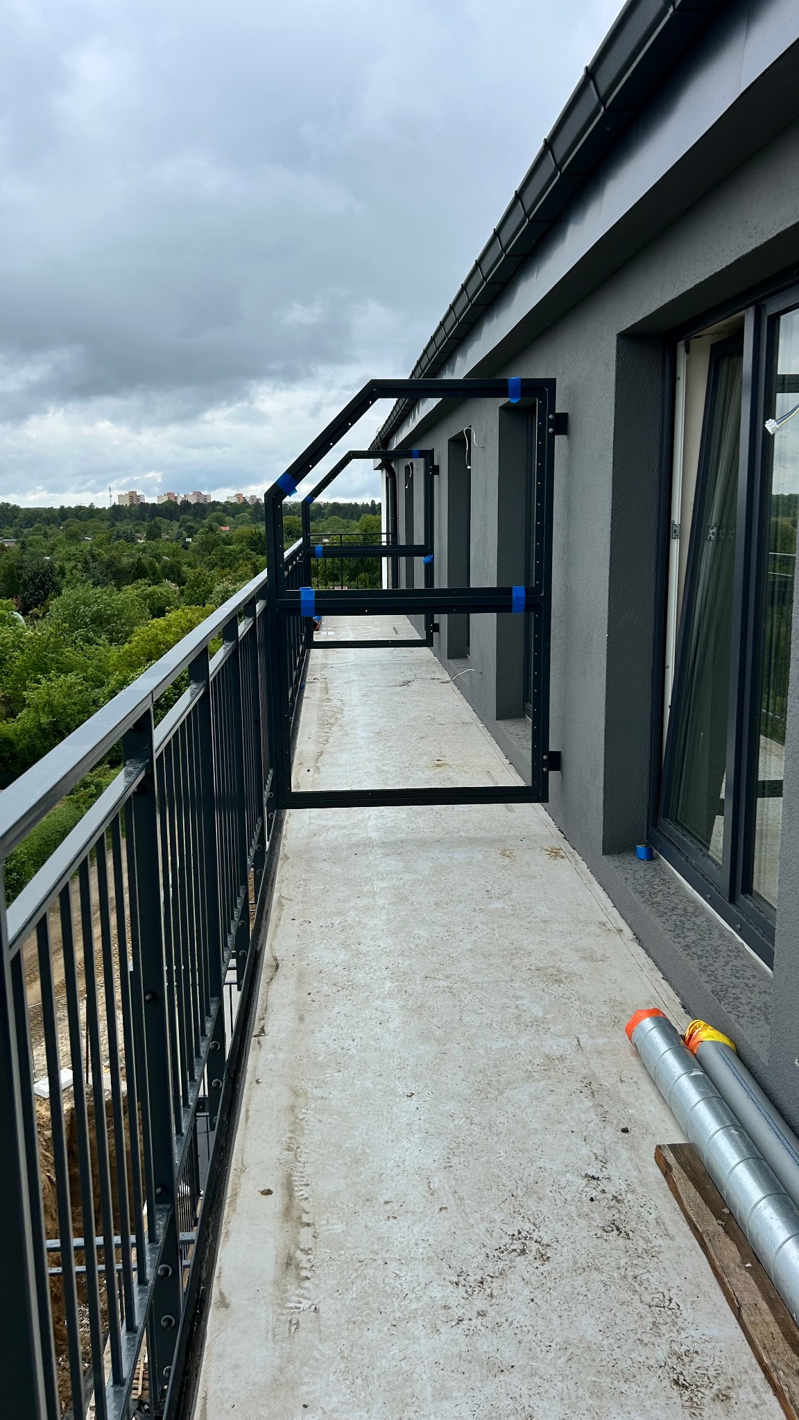 Balkon z zamontowaną, metalową konstrukcją pod balustradę, zabezpieczoną niebieską taśmą. Widoczne elementy montażowe i rury na podłodze balkonu.