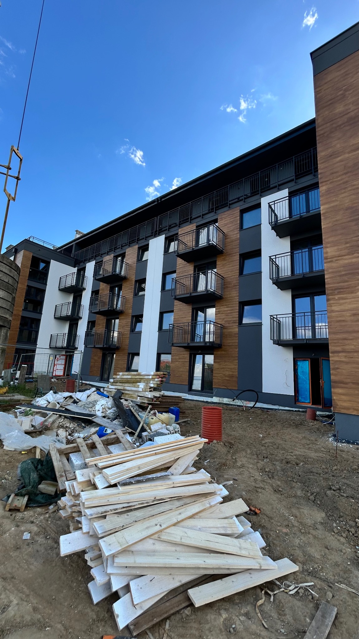 Nowoczesny budynek wielorodzinny z balkonami o metalowych balustradach, w trakcie wykańczania. Na pierwszym planie składowane drewniane deski i gruz budowlany.
