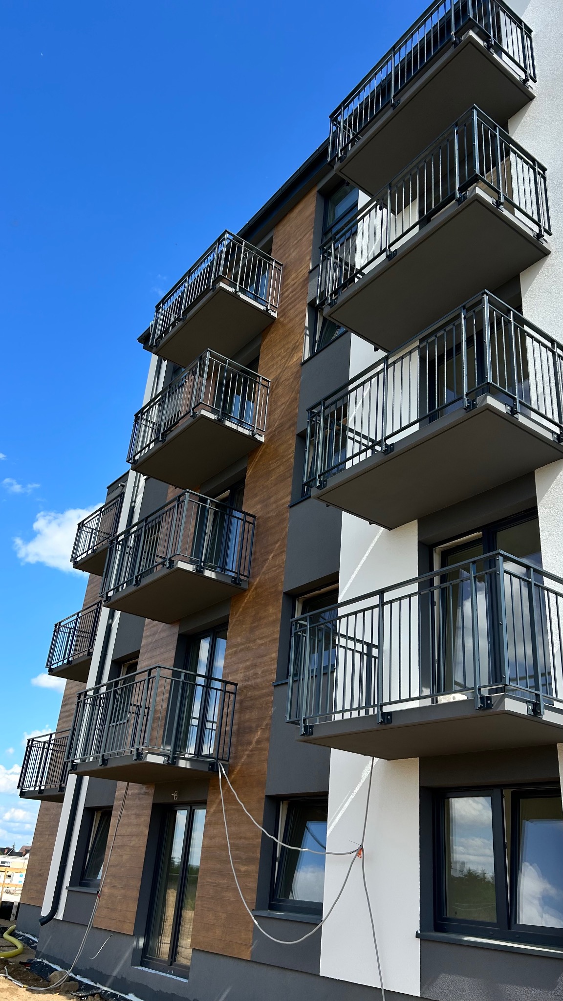 Nowoczesny budynek wielorodzinny z rzędem balkonów z czarnymi, metalowymi balustradami na tle błękitnego nieba. Widok z dołu, podkreślający geometrię i powtarzalność elementów.