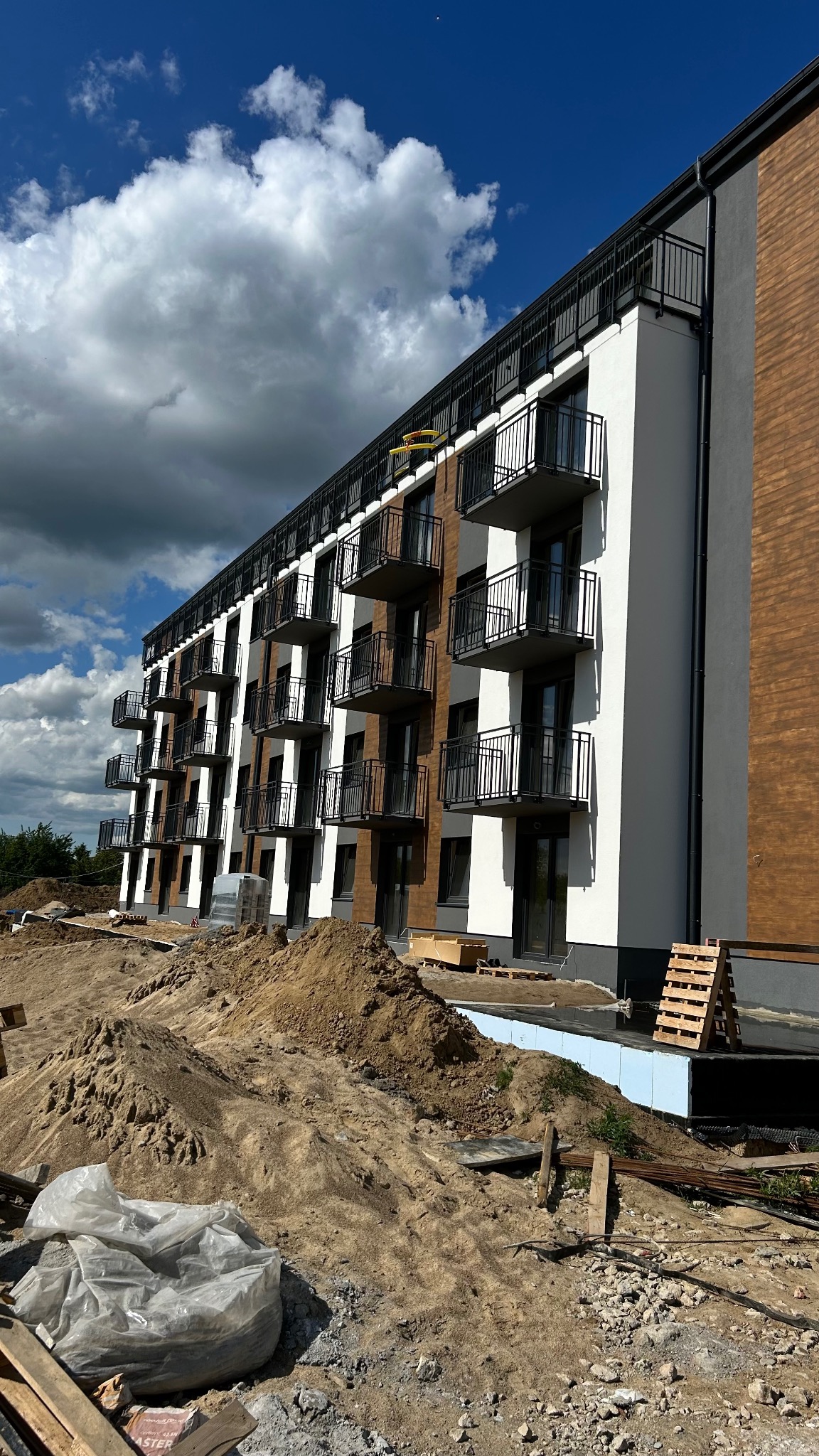 Nowoczesny budynek wielorodzinny z balkonami z czarnymi metalowymi balustradami, w trakcie prac wykończeniowych terenu.